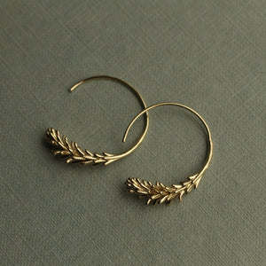 A pair of Tiny Gold Leaf Hoop Earrings displayed on a muted green surface, highlighting the detailed craftsmanship of the rosemary leaf design.