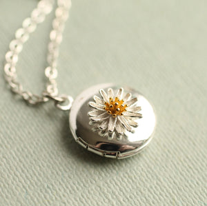 Angled view of miniature silver daisy locket with raised petals and gold centre, displayed on textured pale fabric – dainty botanical pendant.