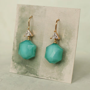 Pair of turquoise hexagon earrings with gold and crystal detail displayed on textured green card, modern geometric earrings.