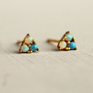 Close-up of gold-plated stud earrings with three opals forming a triangle, glowing with turquoise, gold, and pink tones on a neutral background.