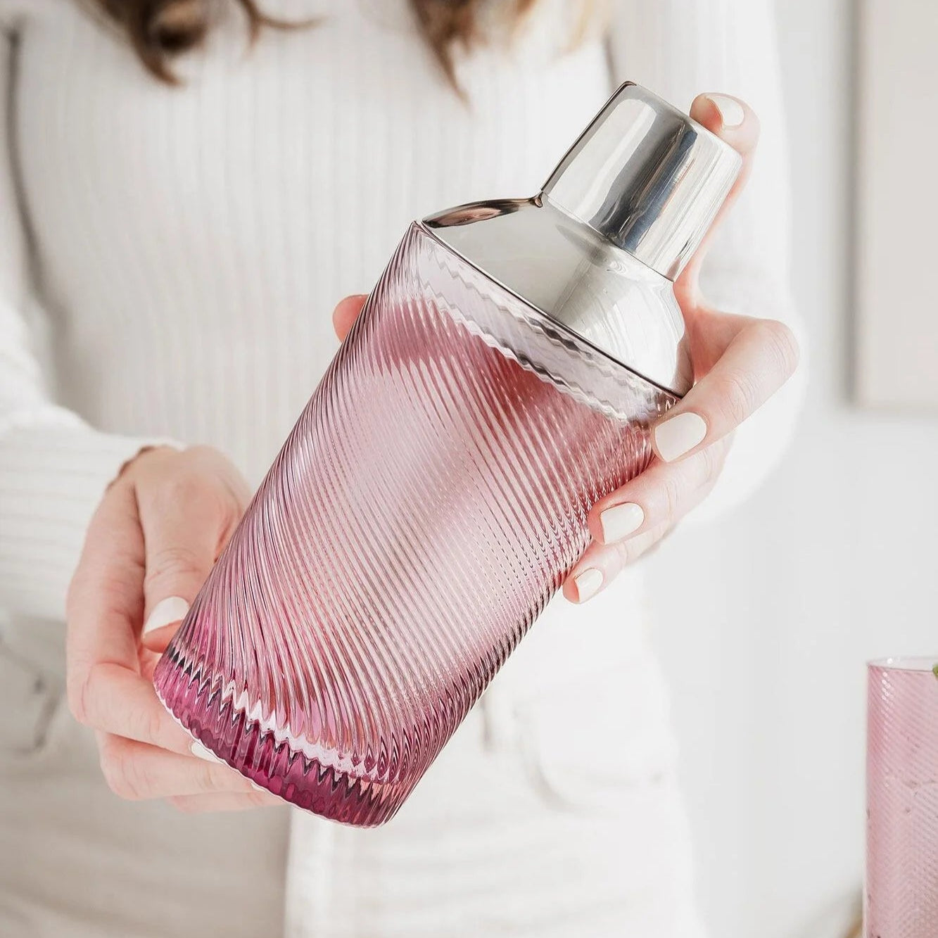 Woman in a white jumper holding the pink twist glass cocktail shaker with both hands, ready to mix a drink.