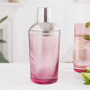 Pink glass cocktail shaker with twisted swirl design and shiny steel top, styled with matching glassware and green leaves.