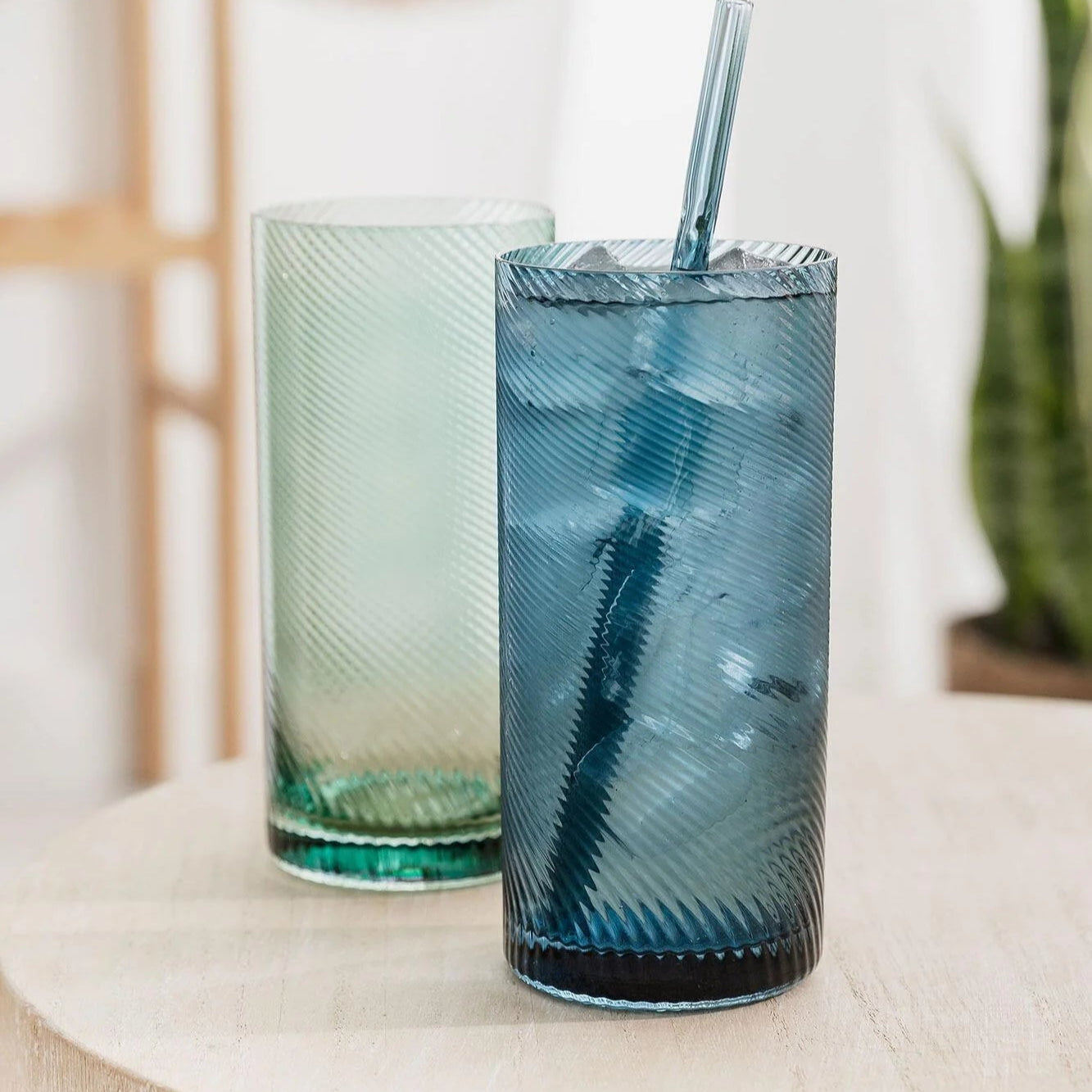Blue and green highball tumblers on a wooden table, with the blue glass filled with ice water and a matching glass straw.