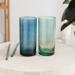 Pair of coloured highball tumblers in blue and green with twisted design, displayed with matching glass straws on a wooden table.