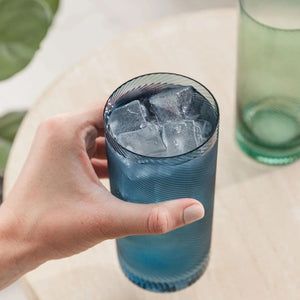 Hand holding the blue coloured highball tumbler filled with ice, green glass tumbler in the background.