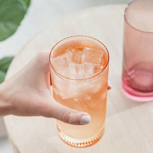 Hand holding peach twist tumbler filled with iced drink next to pink glass.