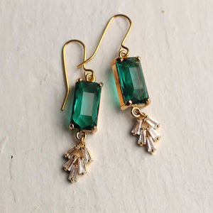 A pair of Vintage Emerald Crystal Chrysler Earrings featuring emerald green baguette-cut glass stones set in brass, with delicate cascading cubic zirconia details. The earrings have gold-plated ear wires and are displayed on a textured white surface.