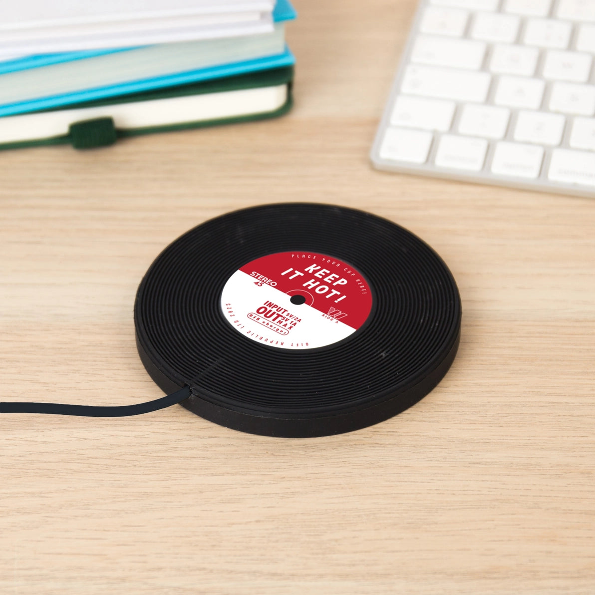 Close-up side view of the Vinyl Mug Warmer shaped like a black vinyl record with red and white centre label reading “Keep It Hot”, shown plugged in on a wooden surface.