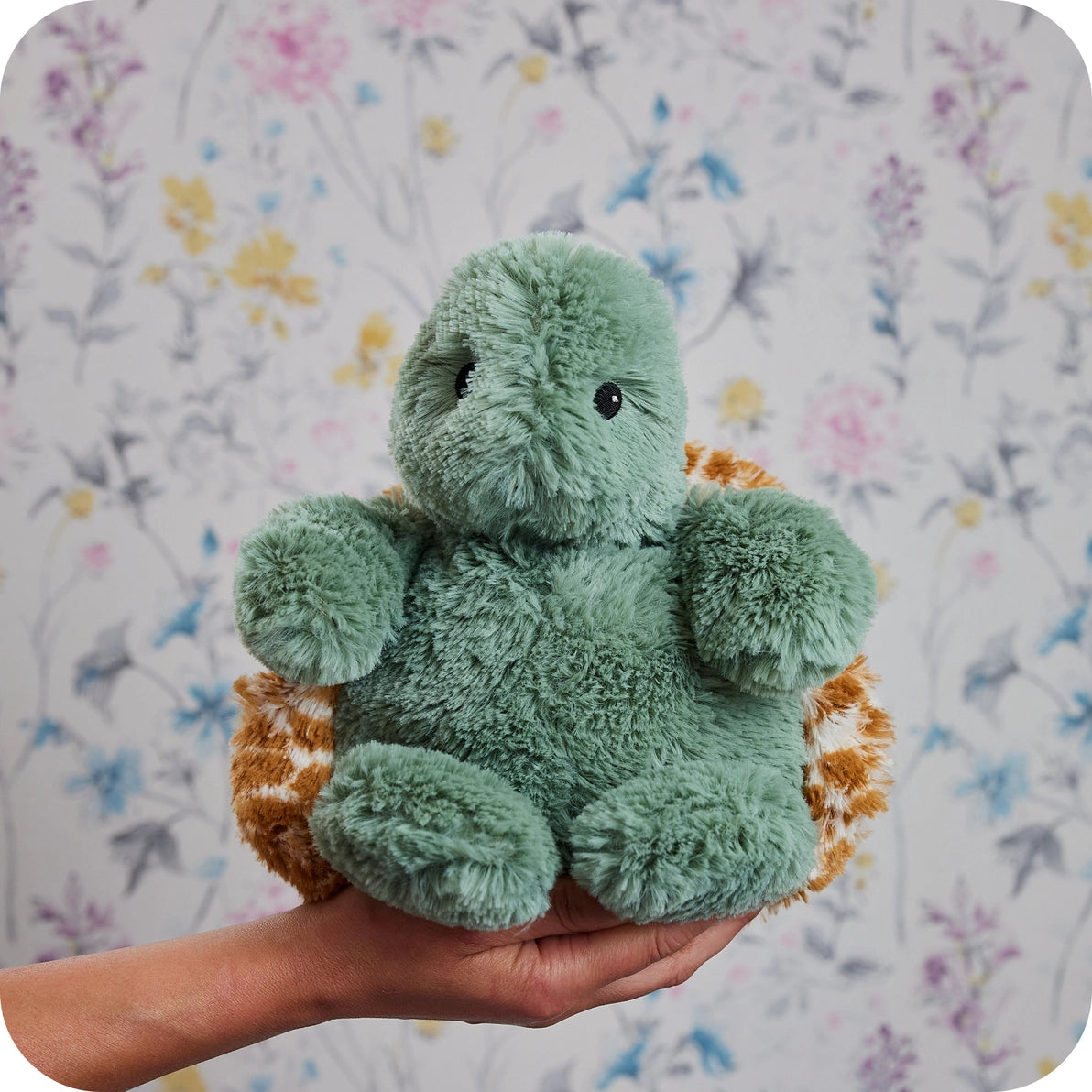 Hand holding Warmies Baby Turtle plush toy against floral background.