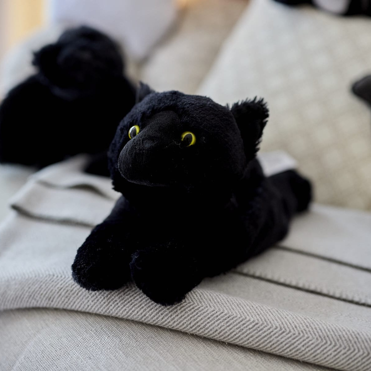 Warmies Black Cat microwavable plush toy resting on a beige sofa throw, creating a warm and cosy home setting.