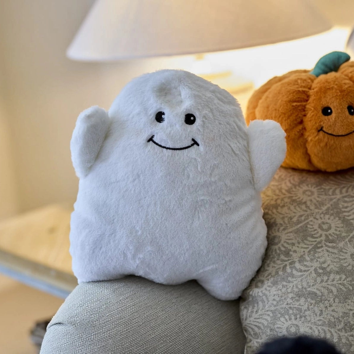 Warmies Ghost plush toy on sofa beside pumpkin cushion, styled in cosy home setting.