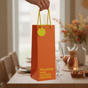 Burnt orange bottle gift bag with yellow warning text held over dining table