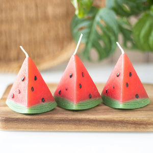 Three watermelon wedge candles displayed on a wooden tray with a tropical background.