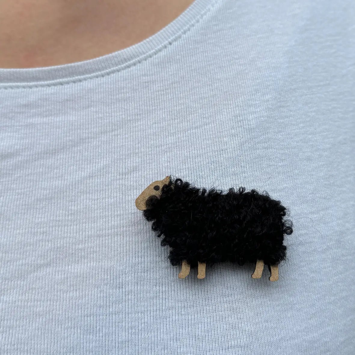 Black Woolly Ewe brooch named Iona pinned to a light shirt, showcasing soft black wool and miniature wooden sheep legs, handcrafted in Scotland.