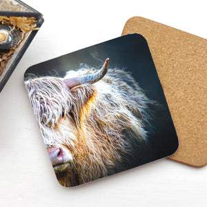 A close-up of the Peekaboo Highland Cow Coaster by Gary Cox, featuring a Highland cow peeking into view. The coaster is made of hardboard with a non-slip cork base.