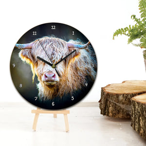 A round wooden wall clock featuring a detailed Highland cow illustration by artist Gary Cox, displayed on a small wooden stand next to a rustic décor setting.