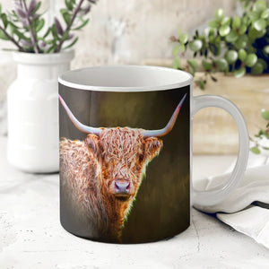 A beautifully printed ceramic mug featuring a Highland cow with long, curved horns against a dark background. The mug is placed on a white countertop with a vase and greenery in the background.