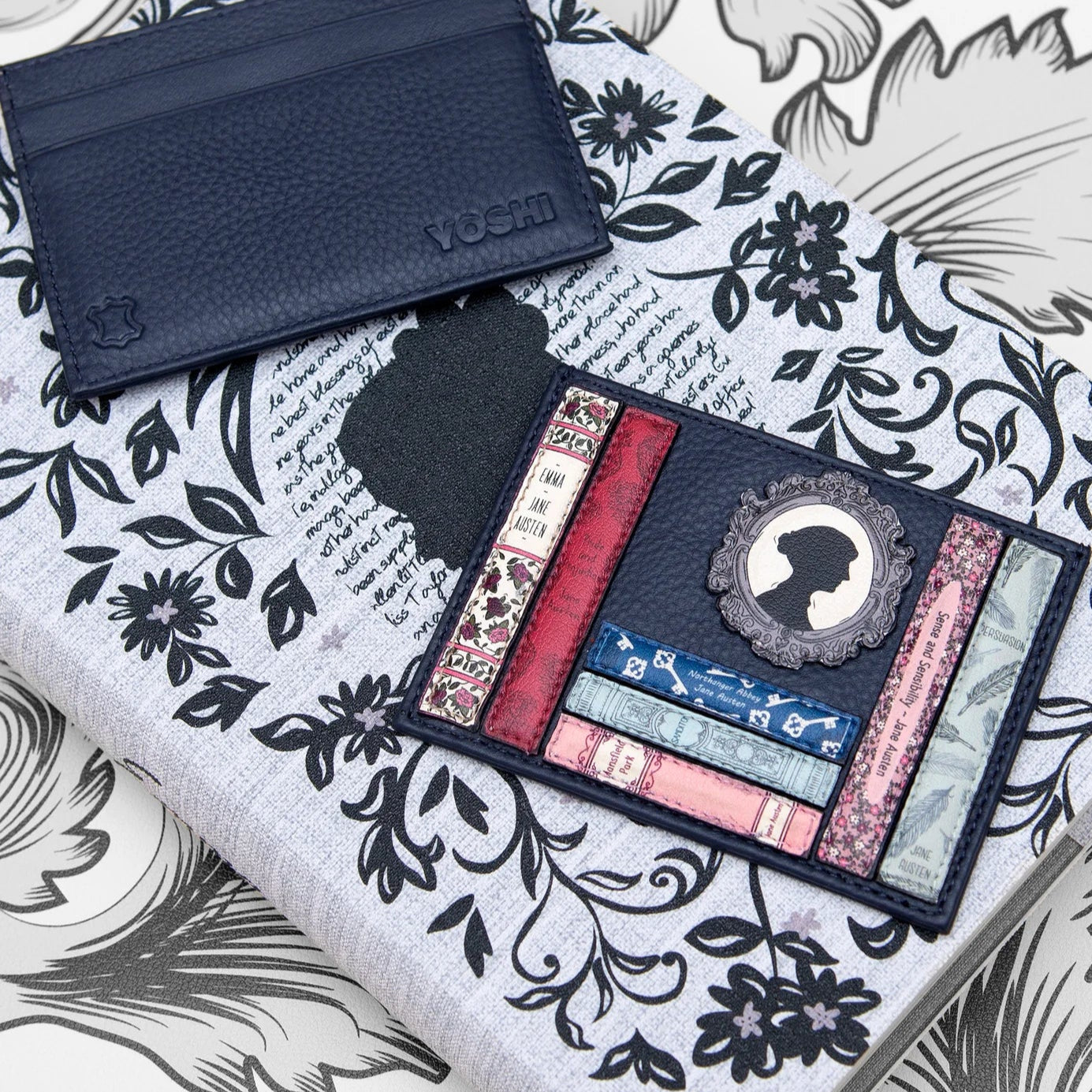 Lifestyle flatlay of Jane Austen Bookworm navy leather card holder, front and back, on floral notebook; compact RFID leather card holder.