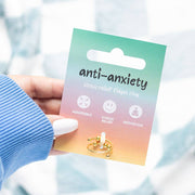 A hand with white nail polish holds up the anti-anxiety fidget ring on its colourful gradient packaging card. The card transitions from turquoise at the top through pink to warm orange at the bottom, displaying the text 'anti-anxiety stress relief fidget ring' along with icons showing adjustable fit, stress relief, and meditation benefits. The gold-toned ring with rotating beads is visible at the bottom of the card. A soft blue knitted sleeve is visible on the arm, with a blurred light background.