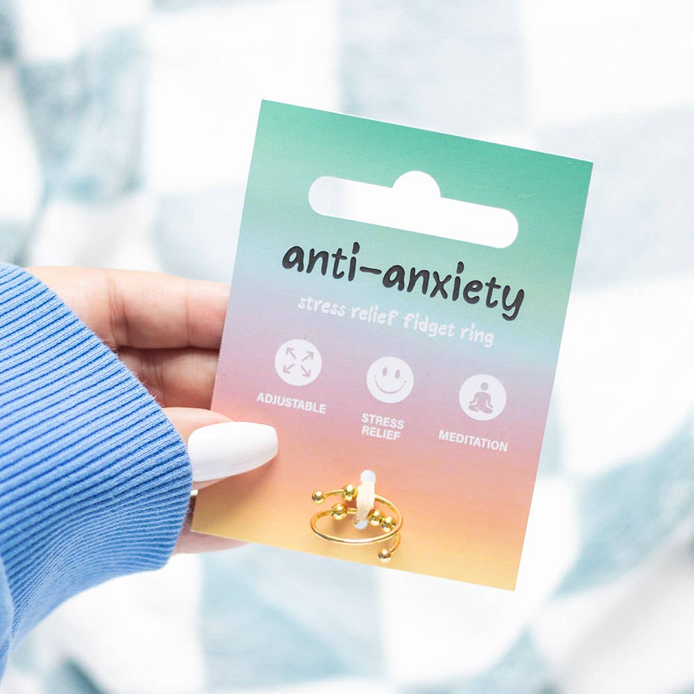 A hand with white nail polish holds up the anti-anxiety fidget ring on its colourful gradient packaging card. The card transitions from turquoise at the top through pink to warm orange at the bottom, displaying the text 'anti-anxiety stress relief fidget ring' along with icons showing adjustable fit, stress relief, and meditation benefits. The gold-toned ring with rotating beads is visible at the bottom of the card. A soft blue knitted sleeve is visible on the arm, with a blurred light background.