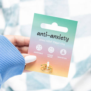A hand with white nail polish holds up the anti-anxiety fidget ring on its colourful gradient packaging card. The card transitions from turquoise at the top through pink to warm orange at the bottom, displaying the text 'anti-anxiety stress relief fidget ring' along with icons showing adjustable fit, stress relief, and meditation benefits. The gold-toned ring with rotating beads is visible at the bottom of the card. A soft blue knitted sleeve is visible on the arm, with a blurred light background.