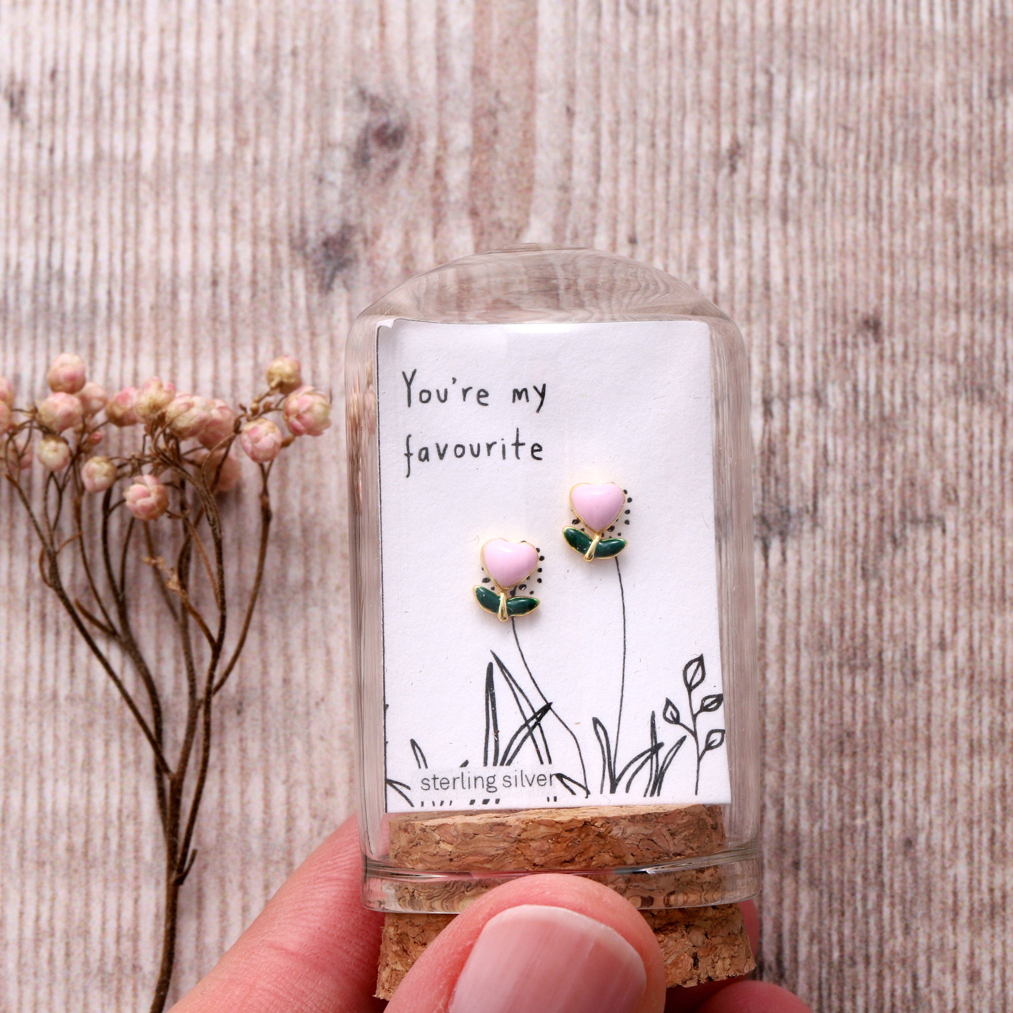Pink enamel flower earrings in glass terrarium held in hand on grey wooden background