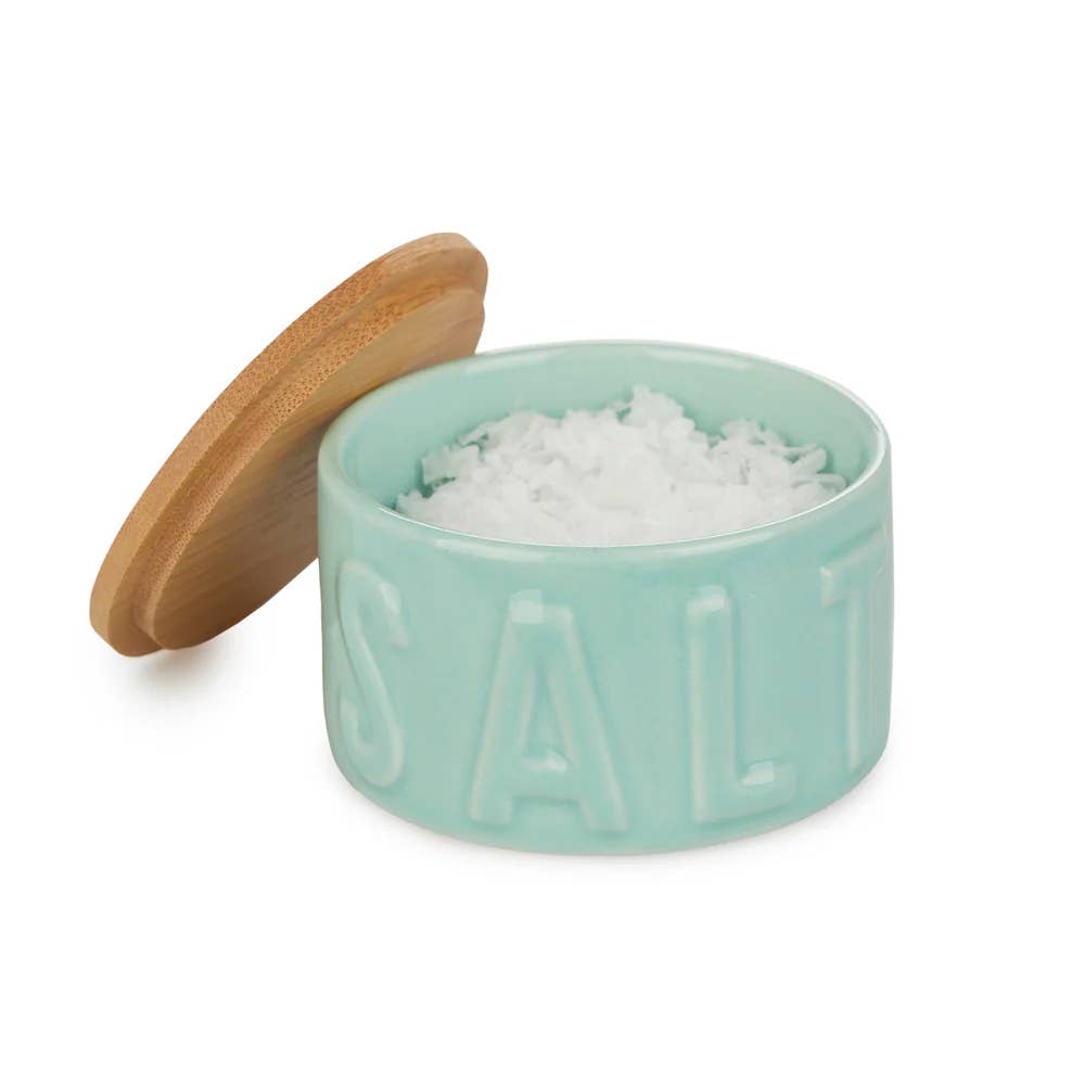 Open turquoise salt cellar filled with salt flakes beside its bamboo lid, white background