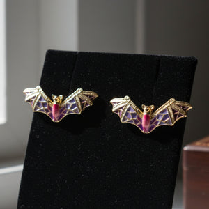 Purple and gold enamel bat stud earrings displayed on black velvet jewelry stand
