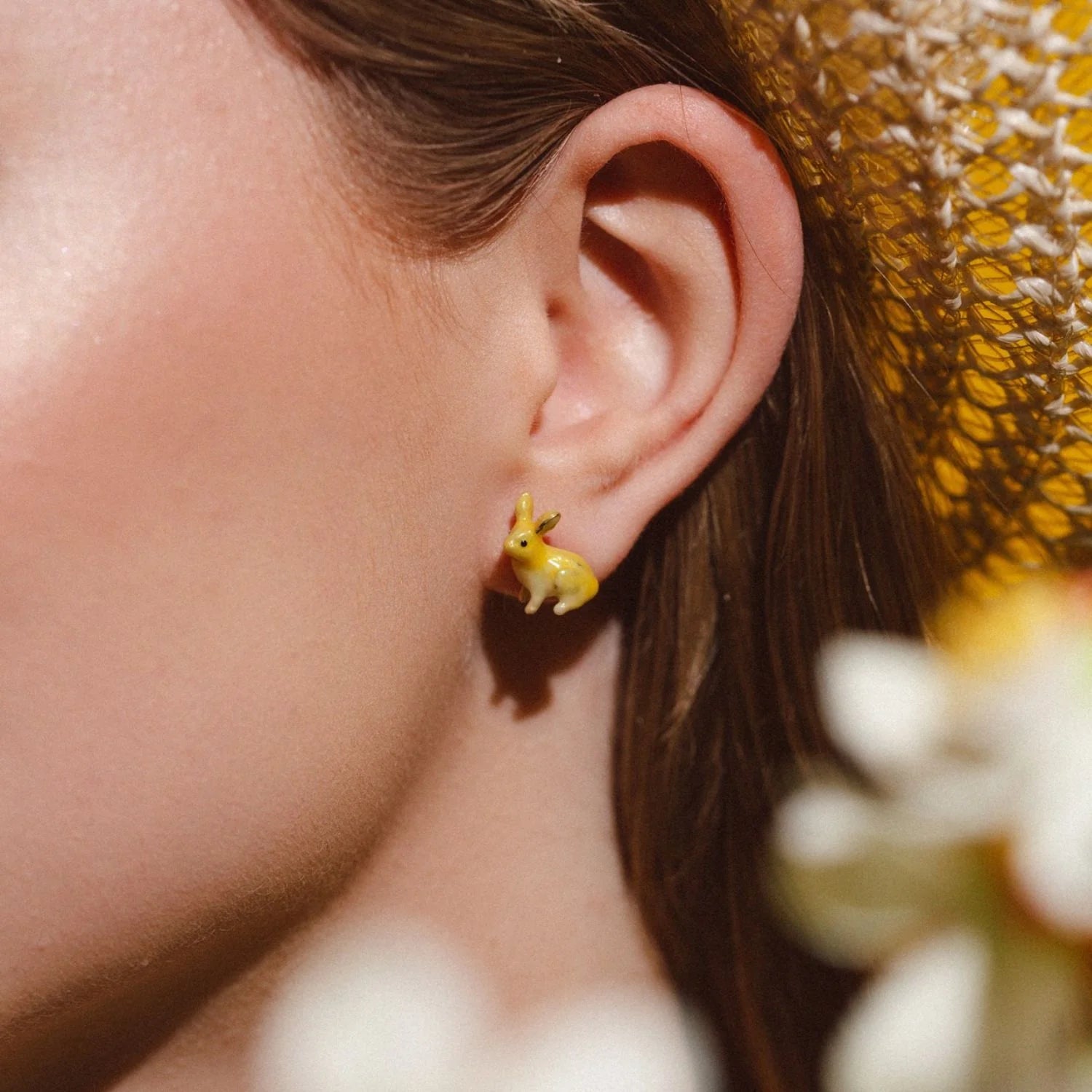 lose-up of woman wearing yellow enamel rabbit stud earring by Bill Skinner, styled with straw hat and soft floral background.