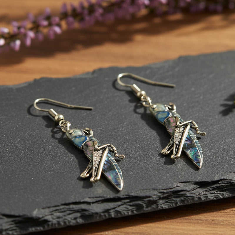 Green resin grasshopper drop earrings with crystal accents displayed on dark slate with purple heather flowers