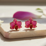 Pink orchid stud earrings with gold detailing displayed on cream marble surface with orchid flowers in background