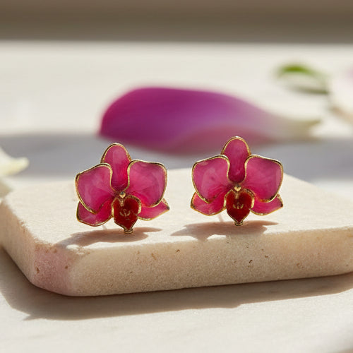 Pink orchid stud earrings with gold detailing displayed on cream marble surface with orchid flowers in background