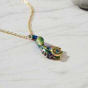 Blue and green enamel peacock pendant necklace with gold chain displayed on light marble surface