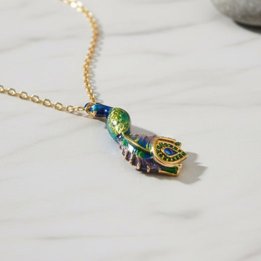 Blue and green enamel peacock pendant necklace with gold chain displayed on light marble surface