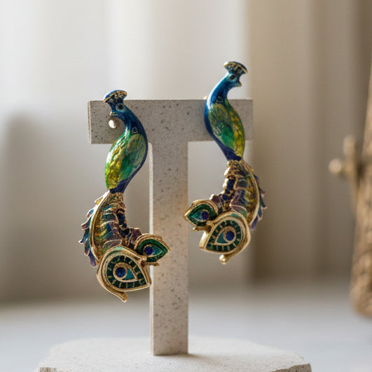 Blue and green enamel peacock stud earrings displayed on gray concrete T-stand