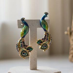 Blue and green enamel peacock stud earrings displayed on gray concrete T-stand