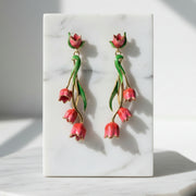 Red tulip drop earrings with gold accents and green enamel leaves displayed on cream marble tile