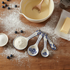 Blue Willow floral ceramic measuring spoons displayed with baking ingredients including flour, blueberries, butter and mixing bowl on wooden surface