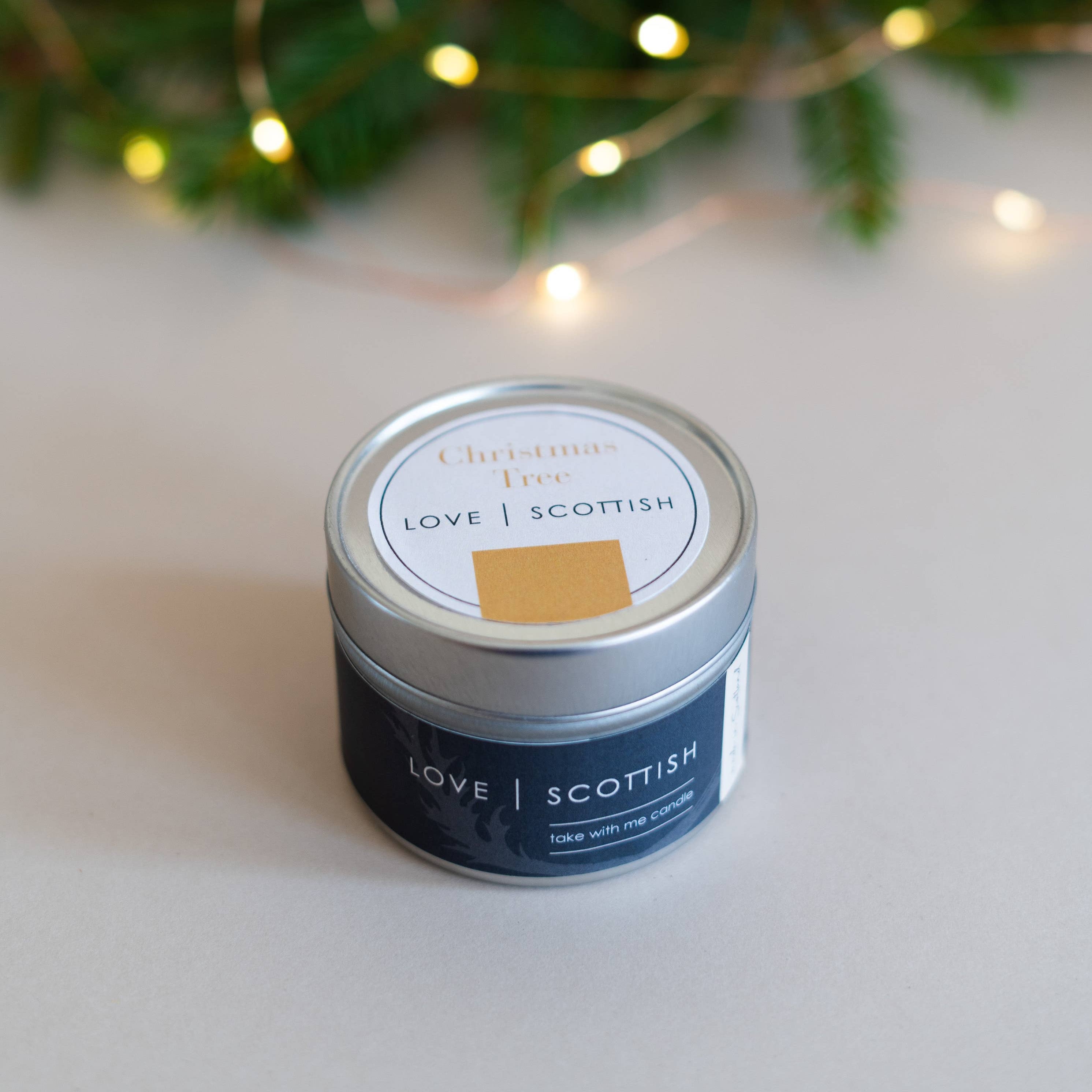 Christmas Tree candle tin with “Love Scottish” label surrounded by pine branches and fairy lights.