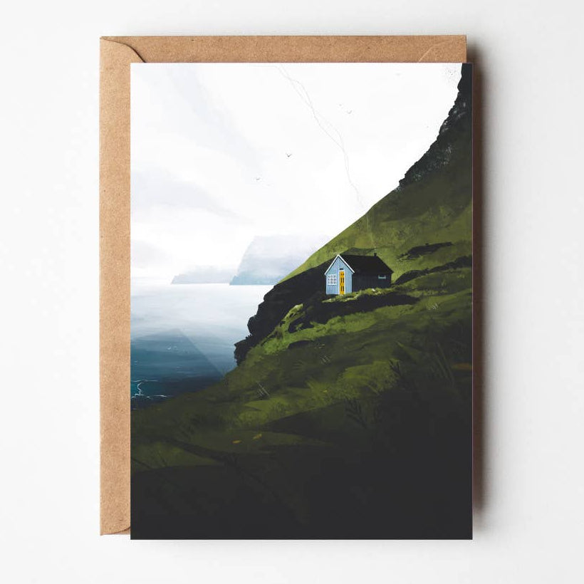 Illustrated Coastal Rise greeting card featuring a cosy cottage on a green Faroe Islands cliffside overlooking the ocean, paired with a kraft envelope.