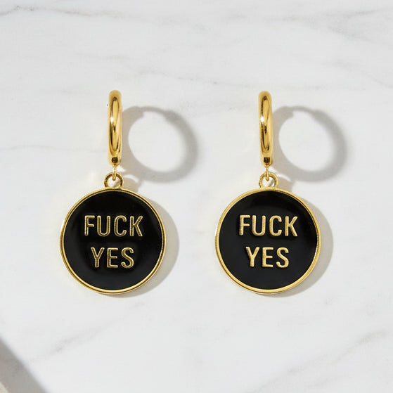 Pair of gold and black hoop earrings that say Fuck Yes against a marble table top. 
