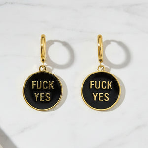 Pair of gold and black hoop earrings that say Fuck Yes against a marble table top. 