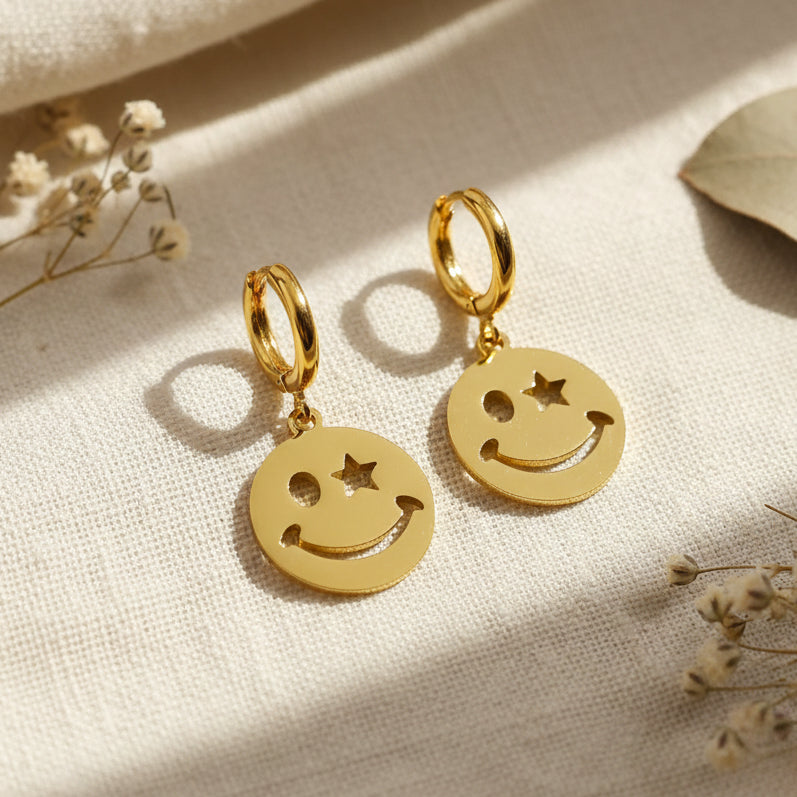 Gold smiley charm earrings displayed on a cloth background. 