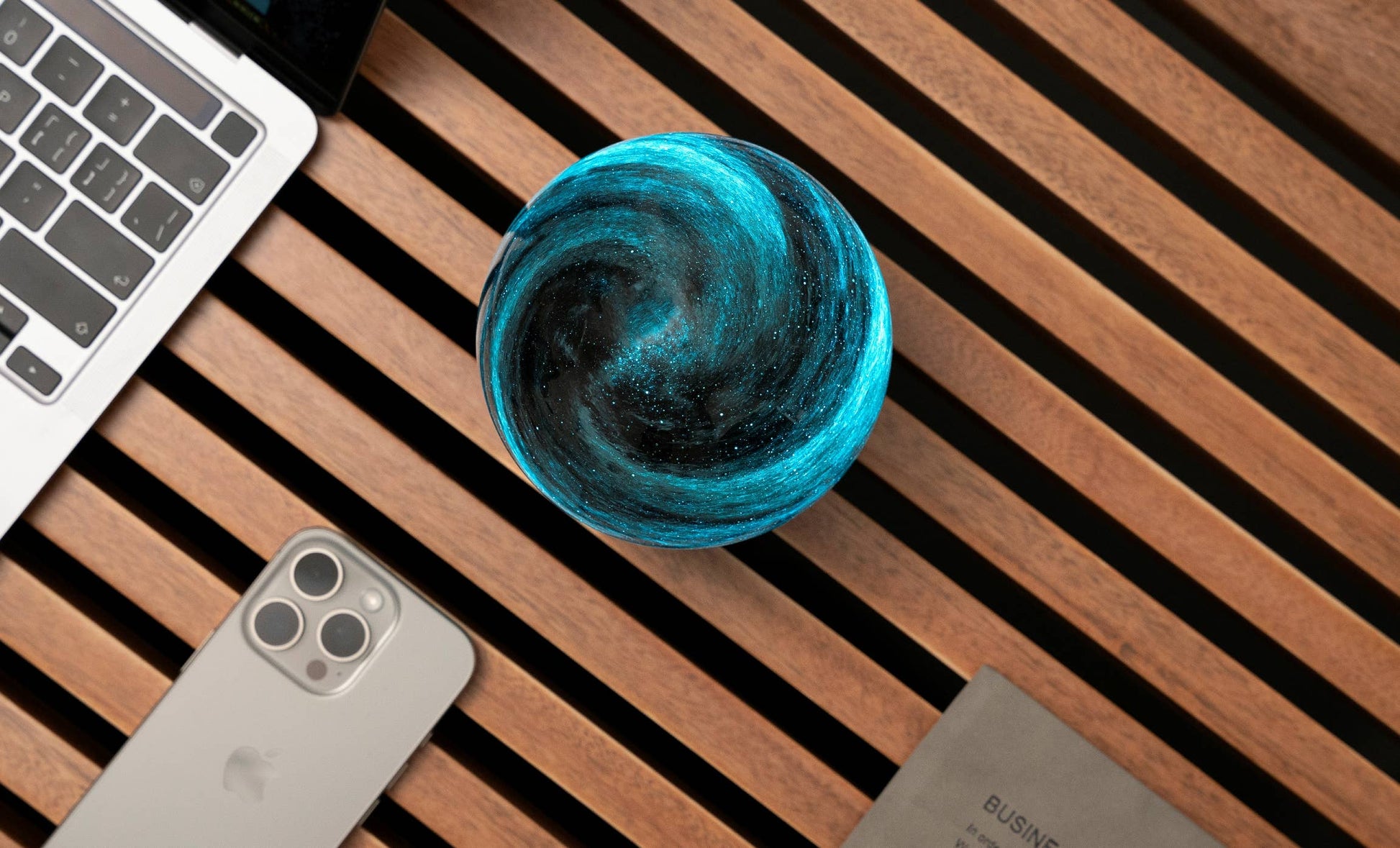 AstraGlass orb overhead view swirling teal and black beside phone