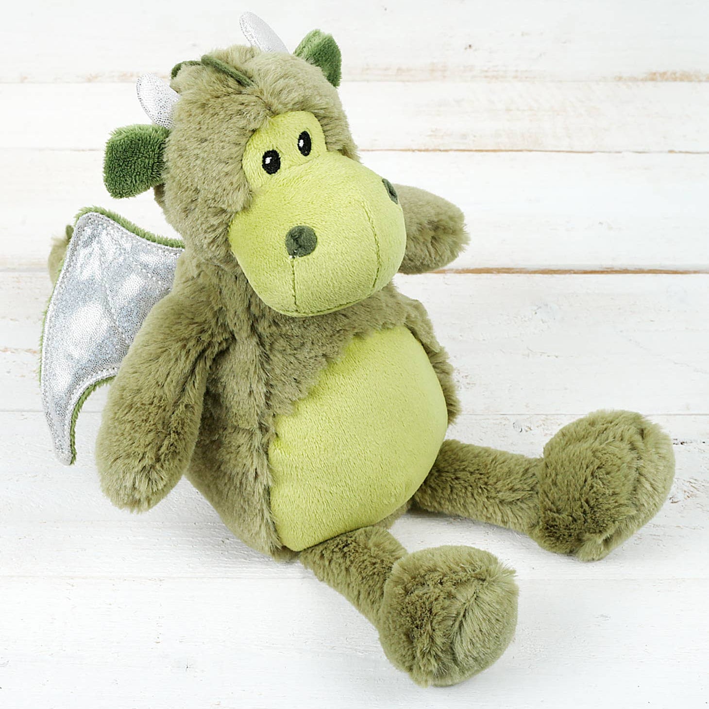 Green dragon plush soft toy with silver wings sitting against a white wooden background