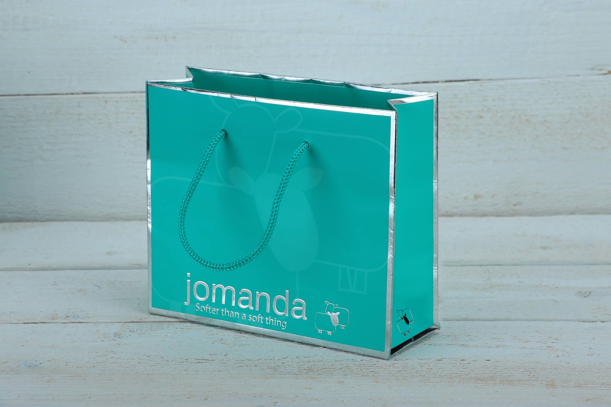 Jomanda teal gift bag with silver edges on a pale blue wooden background