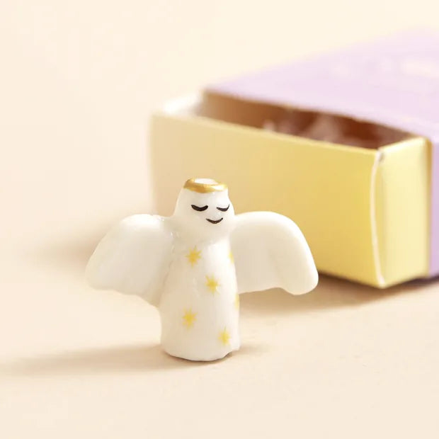 Ceramic angel token with gold halo and star details beside open matchbox on cream background