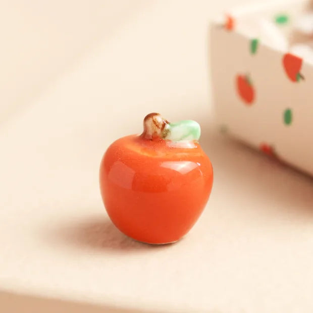 Glossy red ceramic apple token with green leaf beside apple-print matchbox on cream background