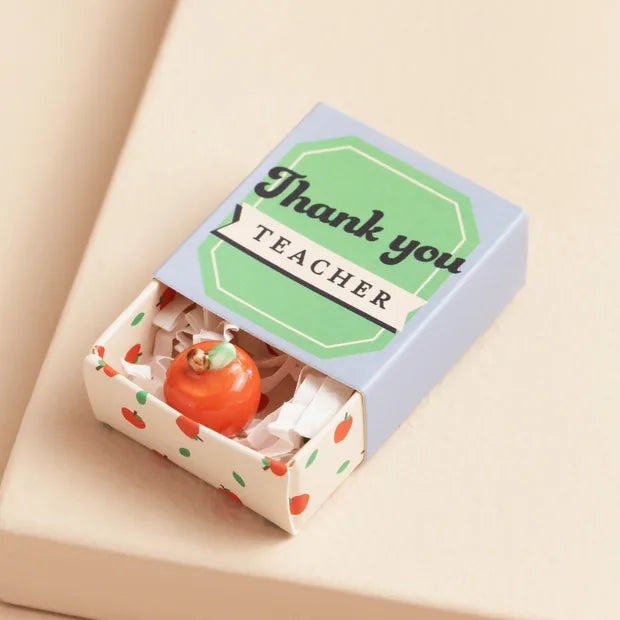 Ceramic apple inside open matchbox with blue and green lid on cream background