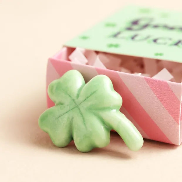 Green ceramic four-leaf clover token beside open pink-striped matchbox on cream background